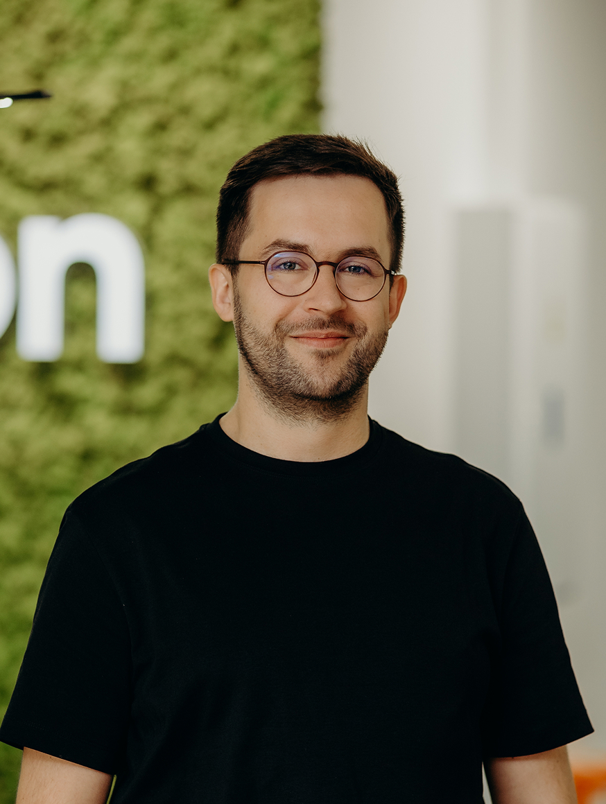 A man standing in the office in front of the Kellton sign, wearing a black shirt and glasses.