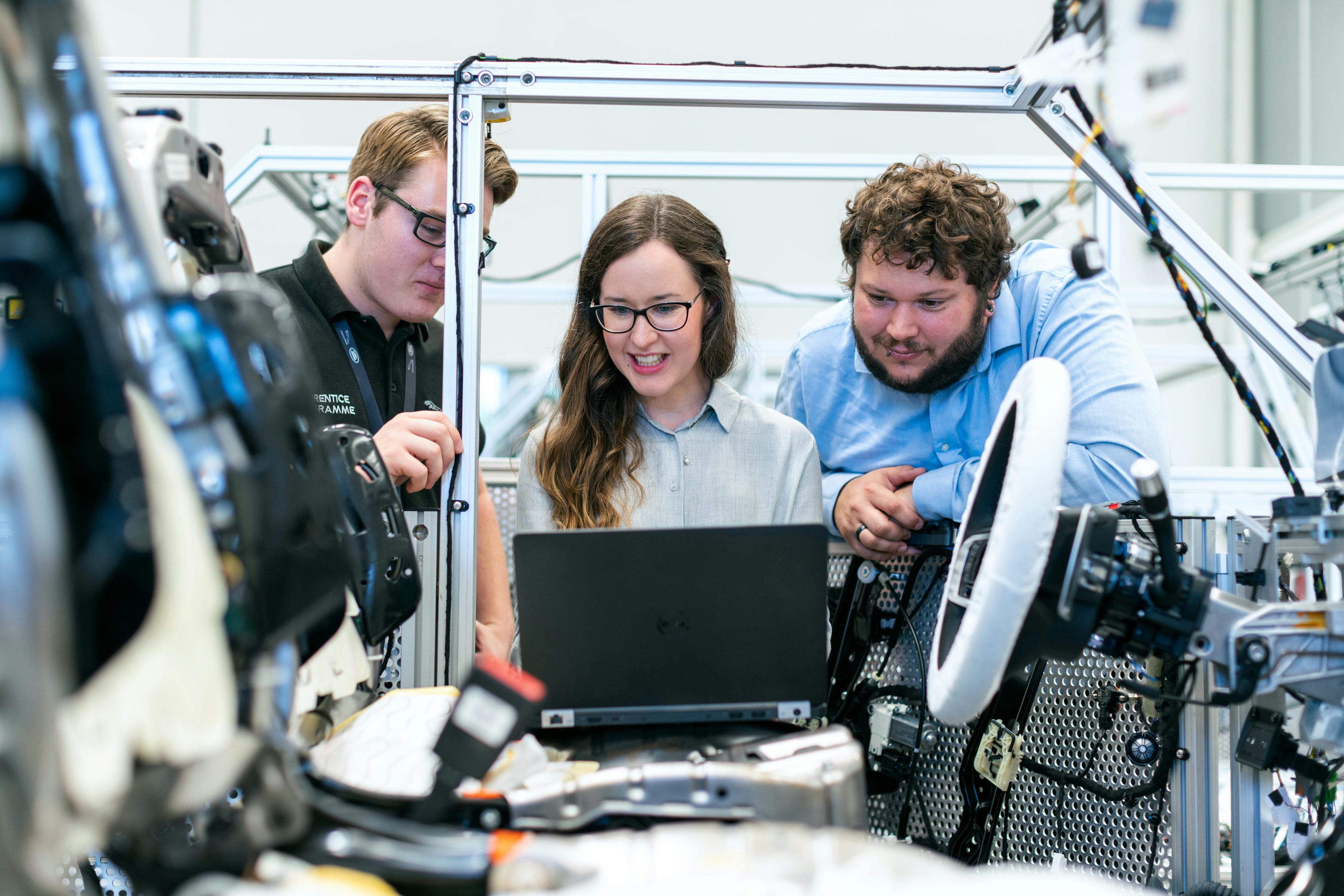 Three people in a robotics factory, the lady in the middle is holding a laptop