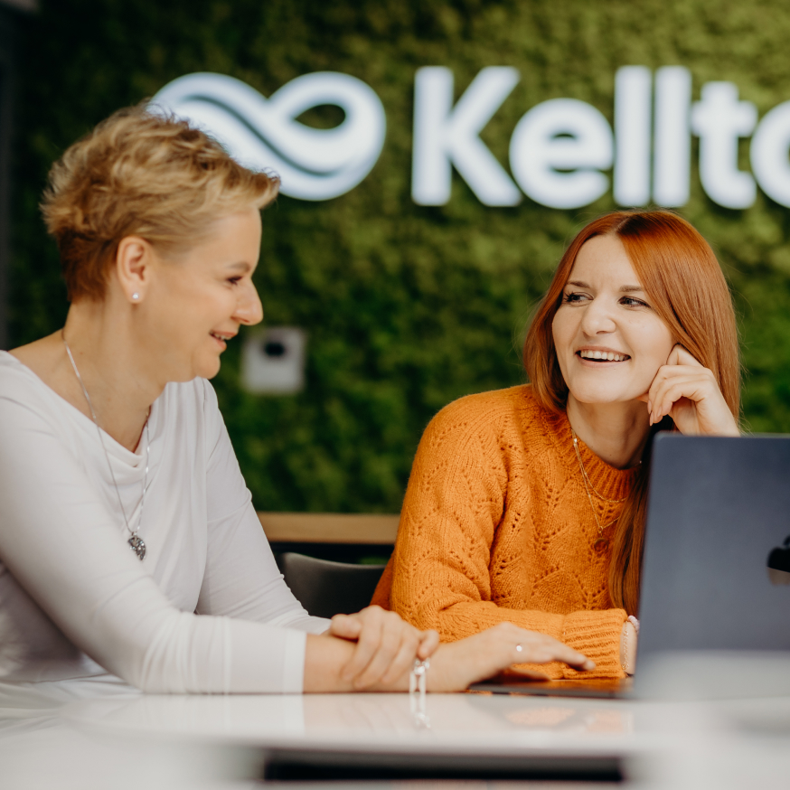 Girls sitting at a table looking at a laptop and smiling. Kellton logo in the background