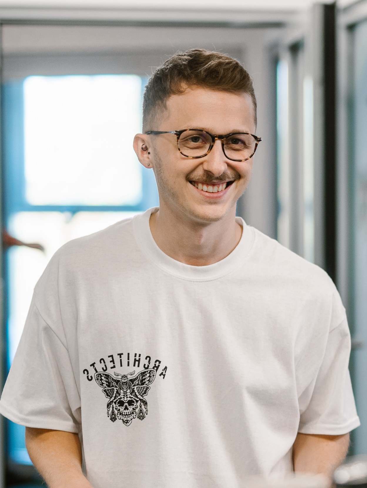 A man standing in the office and smiling, wearing glasses and a white shirt