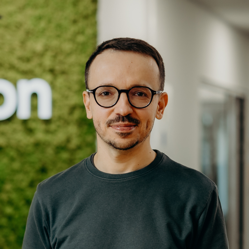 A man standing in the office and smiling, wearing glasses and a green shirt