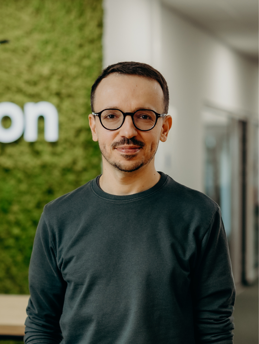 A man standing in the office and smiling, wearing glasses and a green shirt